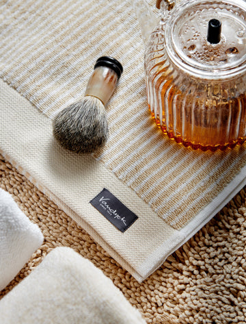 HOME PETIT LIGNE badgoed | bleached sand detail shot van een handdoek in een zacht gele kleur met strepen in het wit, een make-up kwast en een theepot