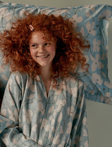 NOA badjas | pearl blue een meisje met oranje haar in een blauwe badjas met witte bloemen en hoofdkussen in lichtblauw met witte bloemen