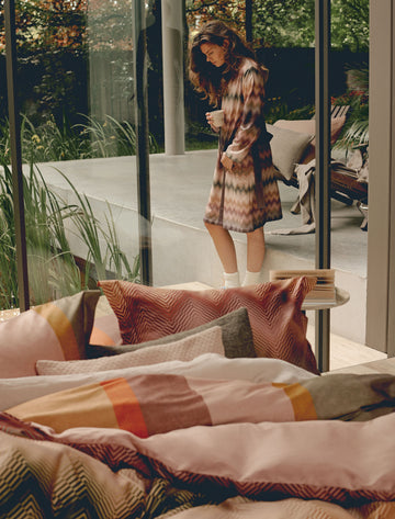 BLURRED LINES dekbedovertrekset | multi Een vrouw in een badjas met zigzag print die en kopje koffie drinkt en een dekbedovertrek in roze en rode kleuren