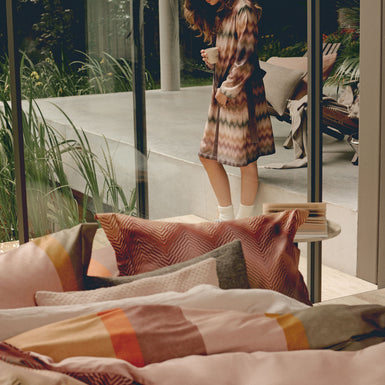 BLURRED LINES dekbedovertrekset | multi Een vrouw in een badjas met zigzag print die en kopje koffie drinkt en een dekbedovertrek in roze en rode kleuren