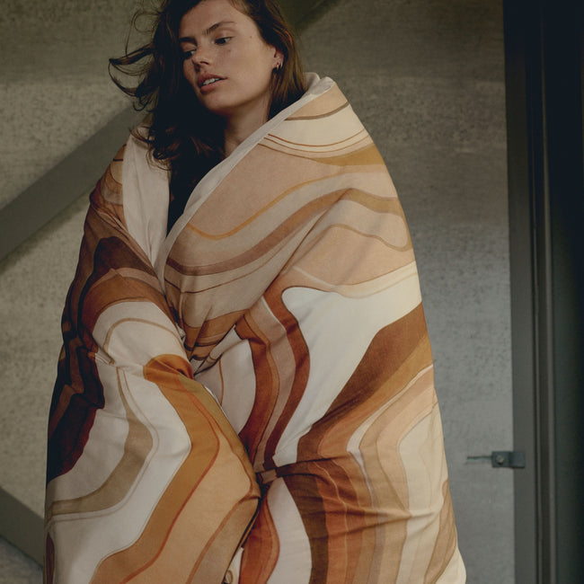 SPIRIT dekbedovertrekset | multi Een vrouw met een dekbedovertrek om haar heen geslagen in een print met bruine en oranje kleuren