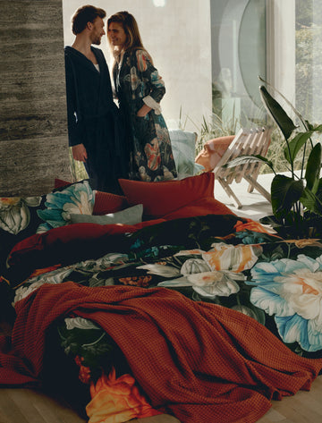 WONDER NIGHT dekbedovertrekset | blue ink Een vrouw en man die achter een bed staan met een bed met dekbedovertrek met bloemenprint en een plaid in een roest bruine/oranje kleur