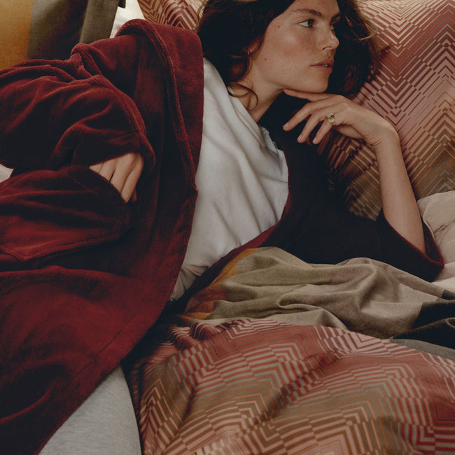 REVEAL dekbedovertrekset | multi Vrouw die op een bed ligt met een rode badjas aan en een dekbedovertrek in zigzag lijnen in roze en rode kleuren