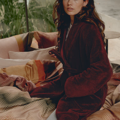 REVEAL dekbedovertrekset | multi Vrouw die op een bed zit met een rode badjas aan en een dekbedovertrek met zigzag strepen en lijnen in roze en rode tinten