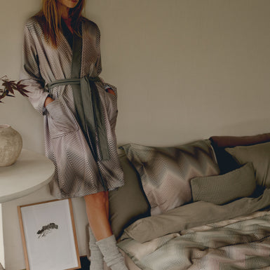 ZIGGY dekbedovertrekset | thyme Een vrouw met een zigzag print badjas aan in groene en paarse kleuren die naast een bed staat met een dekbedovertrek in zigzag print in paarse en groene kleuren