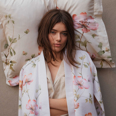 BLOSSOM BREEZE dekbedovertrekset | cream tan Een meid die tegen een muur staat met een kussen achter haar hoofd. Ze draagt een beige badjas met wafelstructuur en een bloemenprint