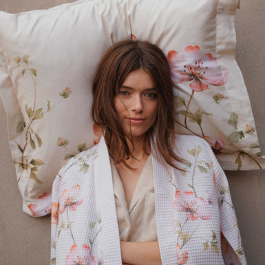 BLOSSOM BREEZE dekbedovertrekset | cream tan Een meid die tegen een muur staat met een kussen achter haar hoofd. Ze draagt een beige badjas met wafelstructuur en een bloemenprint