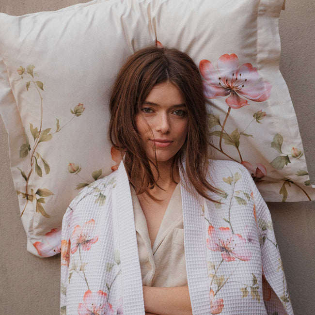 BLOSSOM BREEZE dekbedovertrekset | cream tan Een meid die tegen een muur staat met een kussen achter haar hoofd. Ze draagt een beige badjas met wafelstructuur en een bloemenprint