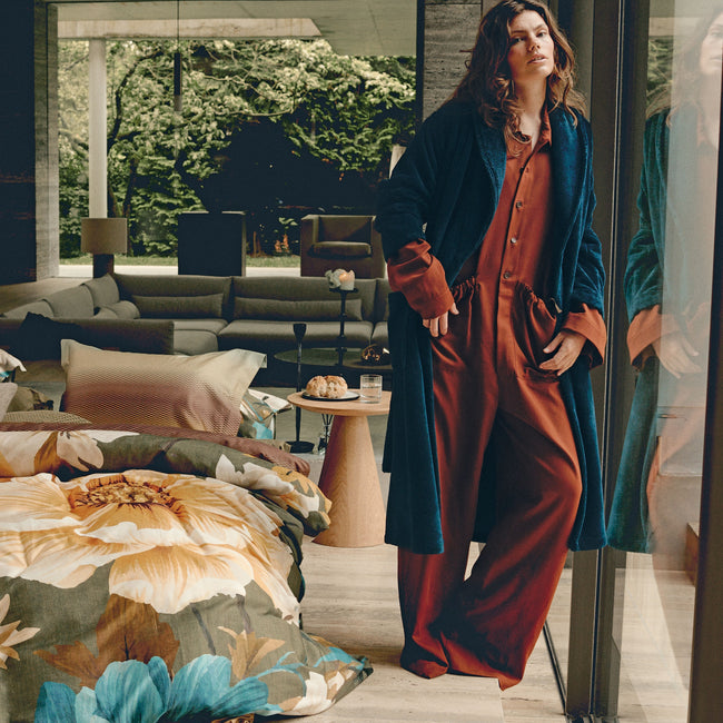 LUMINOUS dekbedovertrekset | multi Een vrouw met een groene badjas aan die naast een bed staat met een dekbedovertrek met bloemen print in gele, blauwe en groene kleuren