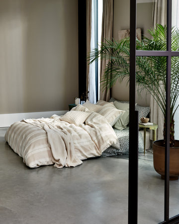 PURE 78 dekbedovertrek | linen Een bed met een overtrek in lichte kleuren met strepen in een mooie en rustige setting met een plant