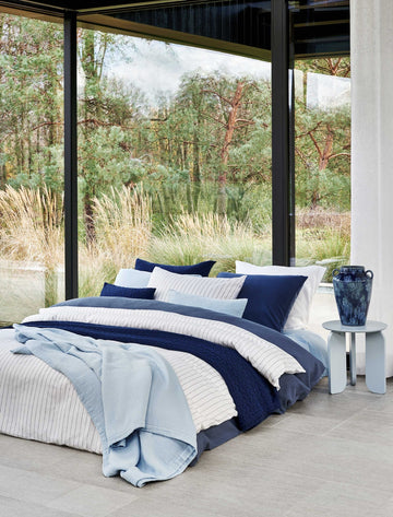 PURE 88 dekbedovertrekset | jeans blue Een bed met een donkerblauw dekbedovertrek, een wit dekbedovertrek met dunne blauwe strepen en op het bed is een lichtblauw plaid gedrapeerd
