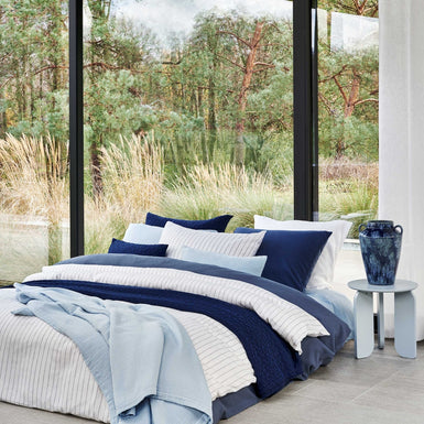 PURE 88 dekbedovertrekset | jeans blue Een bed met een donkerblauw dekbedovertrek, een wit dekbedovertrek met dunne blauwe strepen en op het bed is een lichtblauw plaid gedrapeerd