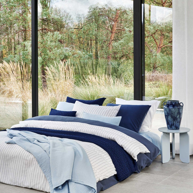 PURE 88 dekbedovertrekset | jeans blue Een bed met een donkerblauw dekbedovertrek, een wit dekbedovertrek met dunne blauwe strepen en op het bed is een lichtblauw plaid gedrapeerd
