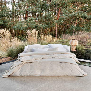 PURITY SORRENTO dekbedovertrekset | white Een bed dat in de buiten ruimte staat. Op het bed ligt een beige dekbedovertrek met een wit patroon en een beige dekbedovertrek met witte strepen. Ook ligt er een beige plaid op het bed