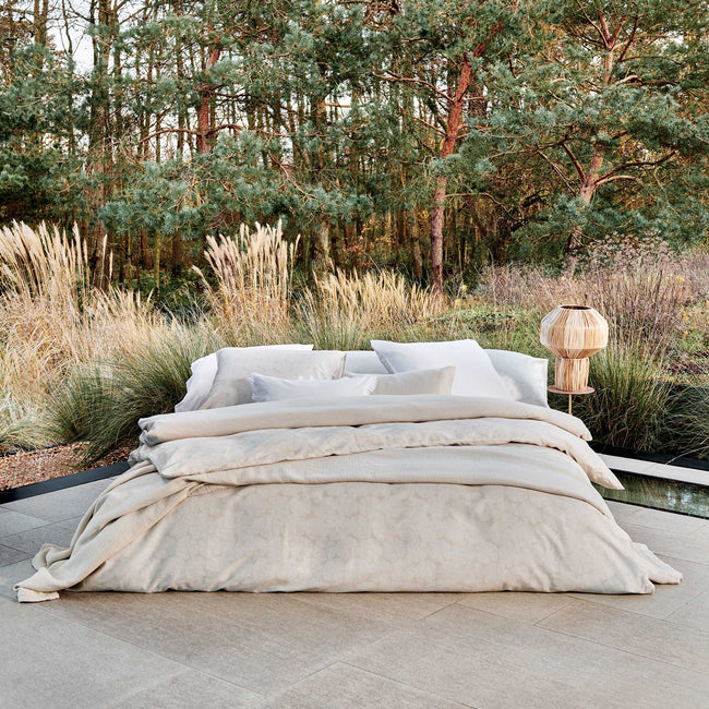 PURITY SORRENTO dekbedovertrekset | white Een bed dat in de buiten ruimte staat. Op het bed ligt een beige dekbedovertrek met een wit patroon en een beige dekbedovertrek met witte strepen. Ook ligt er een beige plaid op het bed