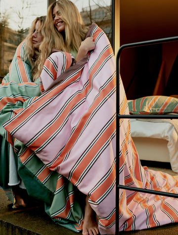 PREPPY STRIPE dekbedovertrekset | smoke green twee meiden met gestreept dekbed om zich heen geslagen in roze en groene tinten, er heerst een gezellige sfeer