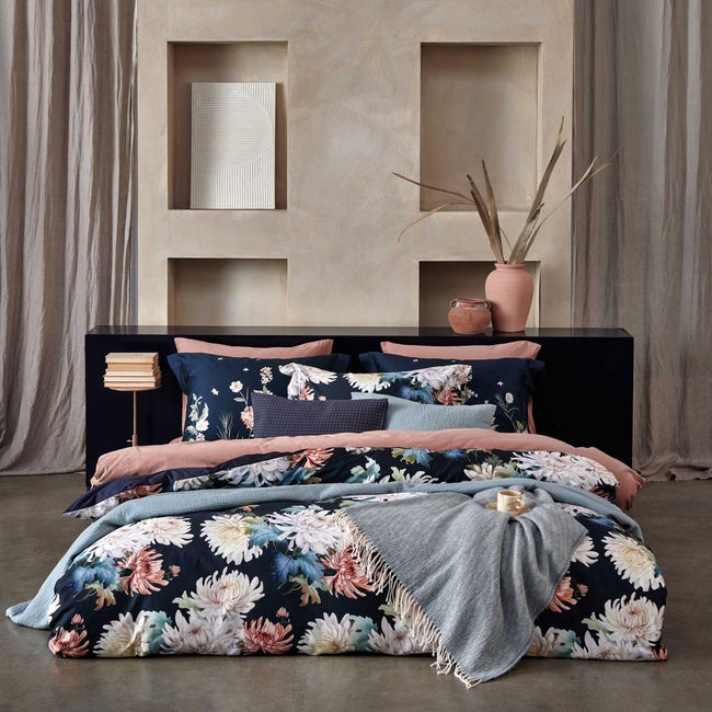 FLORAL BLISS dekbedovertrekset | blue ink een bed in een rustige, serene omgeving met een bed met een dekbedovertrek in donkerblauw met daarop bloemen geprint, een dekbedovertrek in een roze kleur en een sprei van wol en katoen in een blauwe kleur