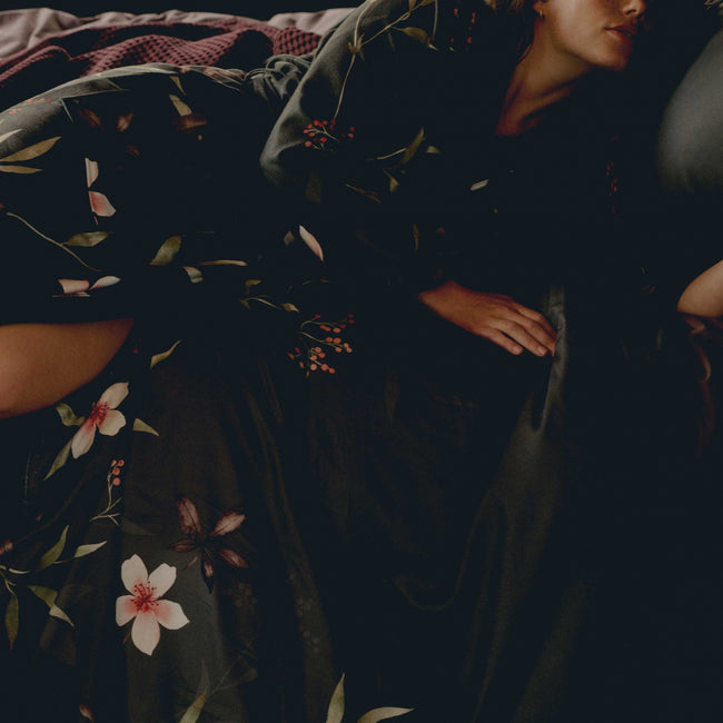 MYSTERY dekbedovertrekset | multi Een vrouw die op een bed ligt met een badjas aan met bloemen in een off black kleur