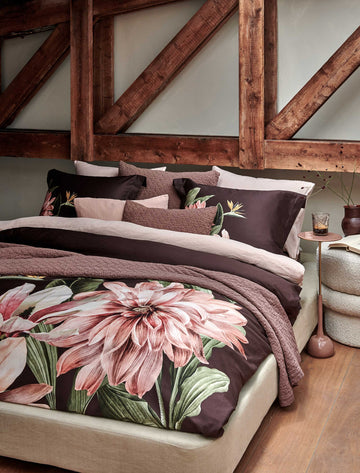 PARADISE BLOOM dekbedovertrekset | dark chocolate Een bed met een dekbedovertrek in een bruine kleur met een grote roze bloem en een sprei en sierkussens in een bruine kleur
