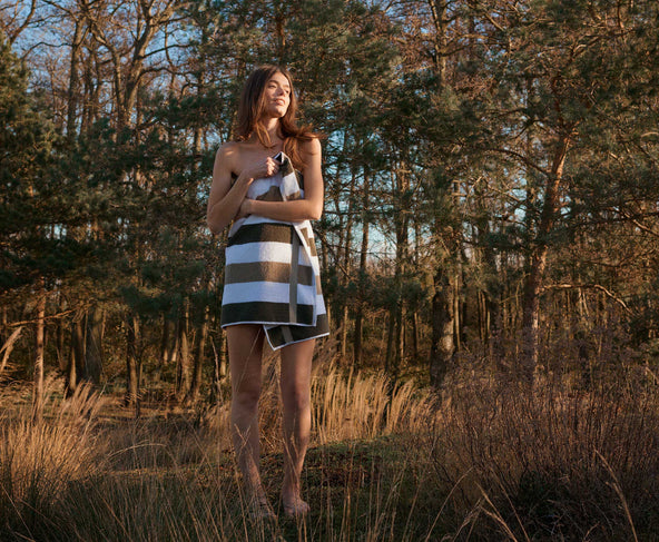 Een vrouw die in de bossen staat met een handdoek in wit met groene strepen om haar lichaam geslagen, het is zonnig weer