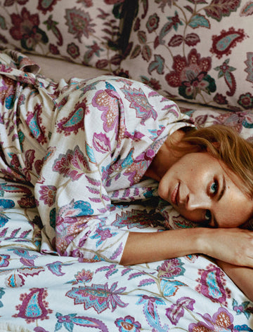 MARY badjas | multi Een vrouw die op een bed ligt met een badjas aan met paisley pirnt in paarse en blauwe kleuren met een dekbedovertrek in dezelfde print