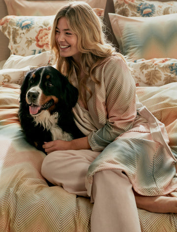 Vrouw op bed met een hond op een bed met een dekbedovertrek met een zig zag print en een badjas aan met een zig zag print