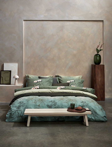 een bed in een rustige, serene omgeving met een dekbedovertrek in groene kleuren met een bloemen print en blaadjes. Voor het bed staat een bankje met daarop een glas en een broodje