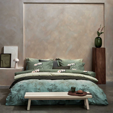 een bed in een rustige, serene omgeving met een dekbedovertrek in groene kleuren met een bloemen print en blaadjes. Voor het bed staat een bankje met daarop een glas en een broodje