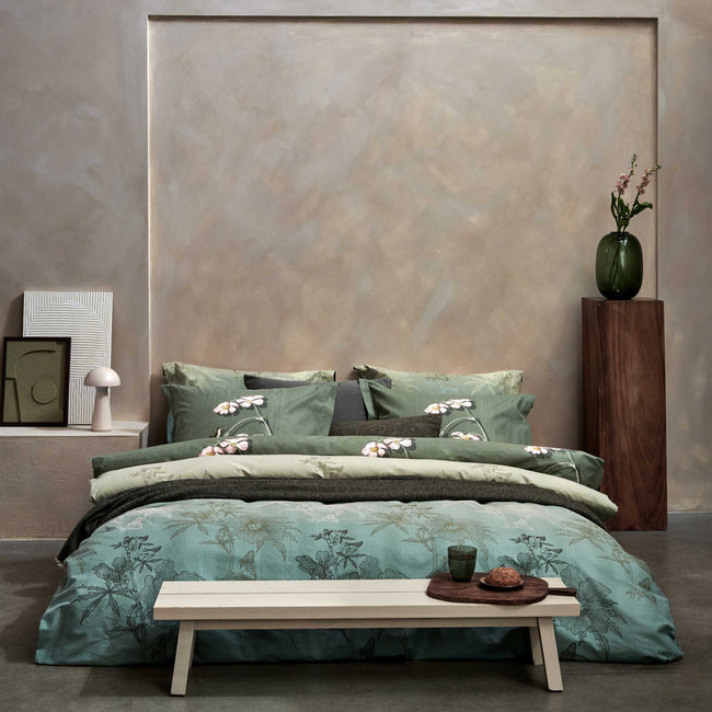 een bed in een rustige, serene omgeving met een dekbedovertrek in groene kleuren met een bloemen print en blaadjes. Voor het bed staat een bankje met daarop een glas en een broodje