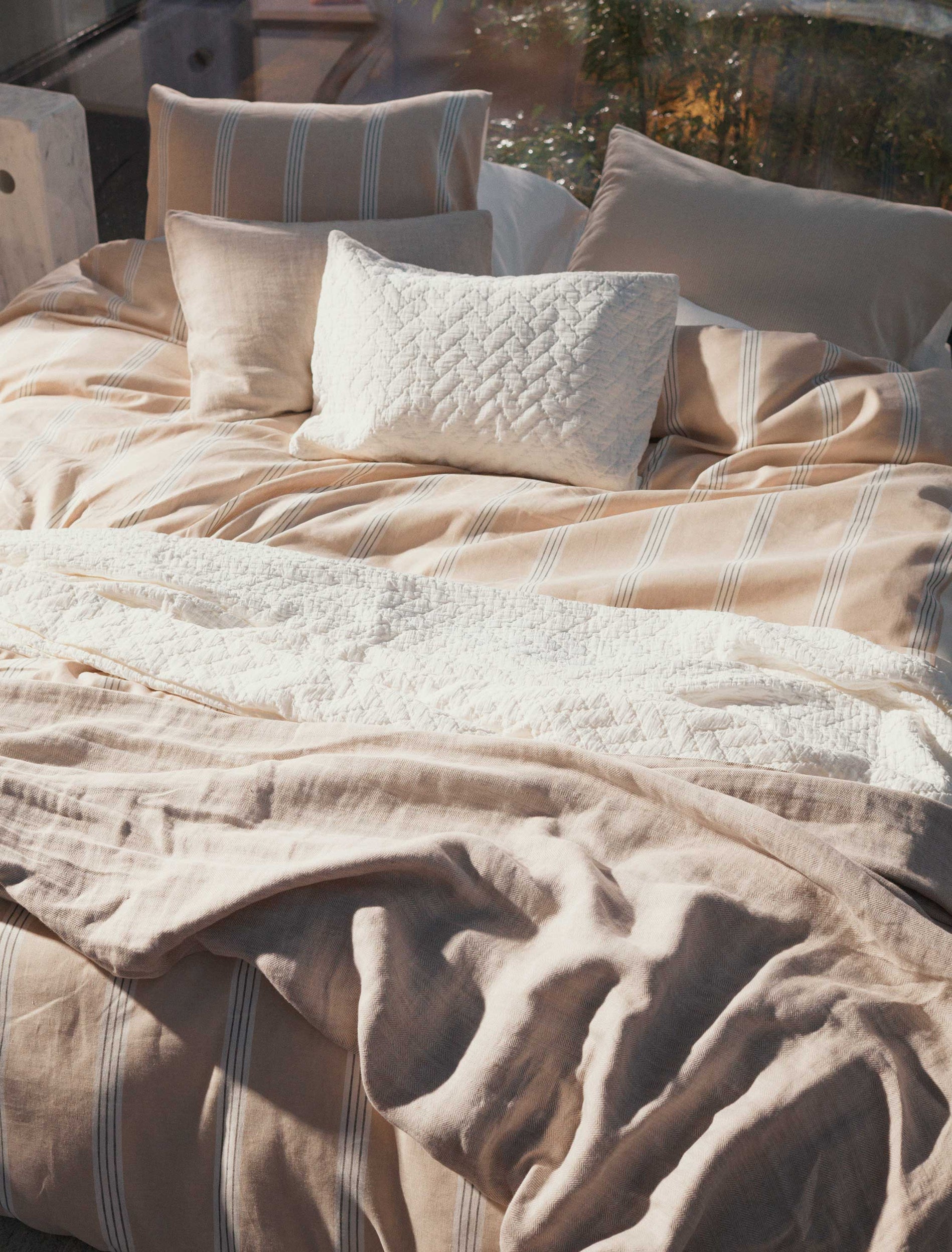 Een bed met een beige dekbedovertrek met witte strepen en een wit plaid op het bed