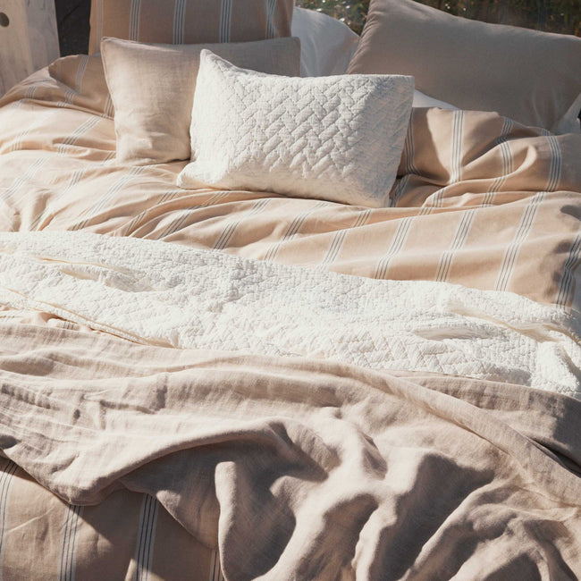Een bed met een beige dekbedovertrek met witte strepen en een wit plaid op het bed