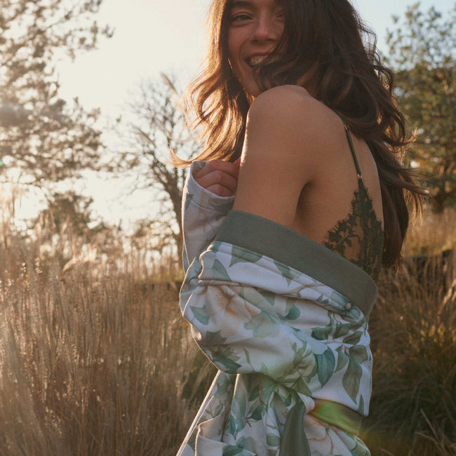 Sofia thyme badjas heeft  grote bloemen met creme en groene tinten met groene bladeren op een off white achtergrond. De basjas heeft grote steekzakken, een brede revers en wordt gesloten met een ceintuur in een lijfgroene tint. 