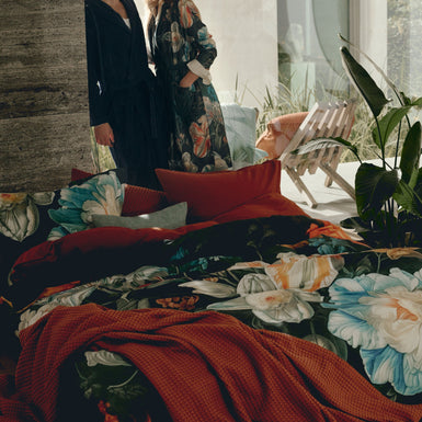 Een vrouw en man die achter een bed staan met een bed met dekbedovertrek met bloemenprint en een plaid in een roest bruine/oranje kleur