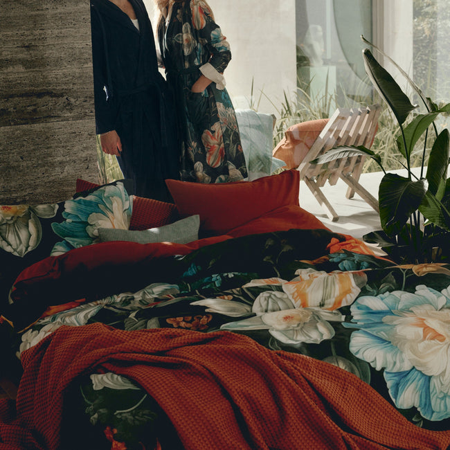 Een vrouw en man die achter een bed staan met een bed met dekbedovertrek met bloemenprint en een plaid in een roest bruine/oranje kleur