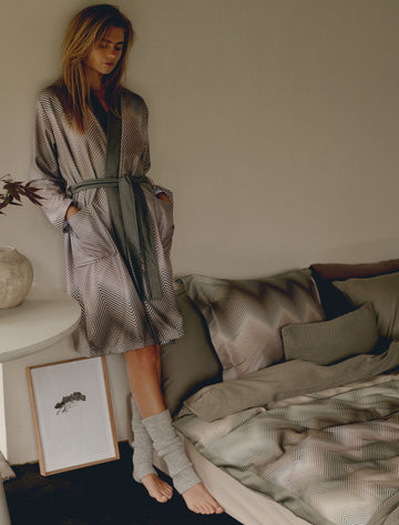Een vrouw met een zigzag print badjas aan in groene en paarse kleuren die naast een bed staat met een dekbedovertrek in zigzag print in paarse en groene kleuren