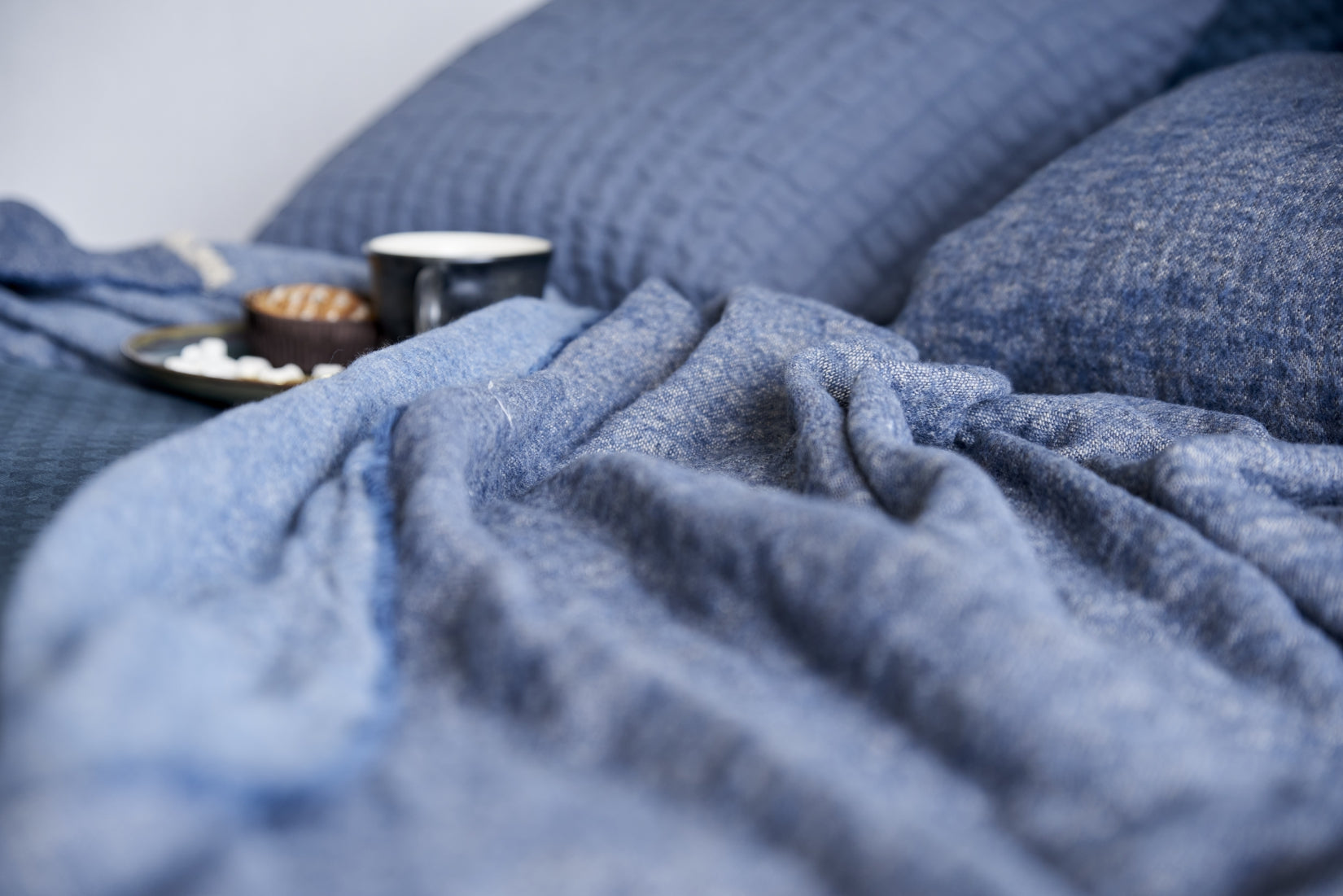 detail shot van een plaid in een jeans blue kleur wat op een bed ligt met een kopje koffie en ontbijt