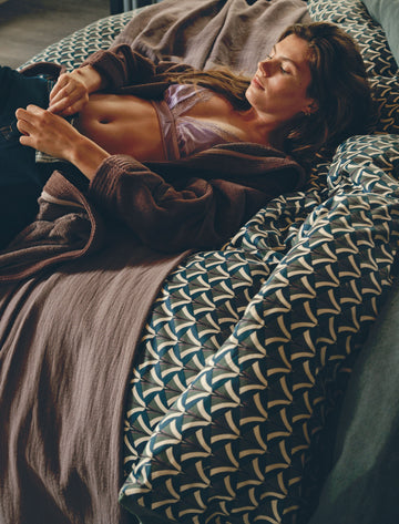 Een meid die op een bed ligt en haar broek vastmaakt met een dekbedovertrek in grafische print in blauwe kleuren en een sprei in een paarse kleur