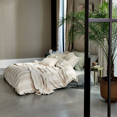 Een bed met een overtrek in lichte kleuren met strepen in een mooie en rustige setting met een plant