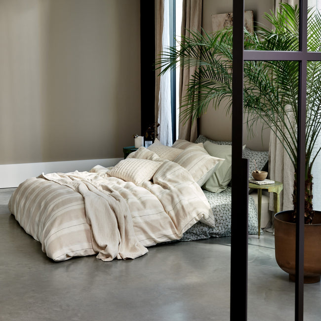 Een bed met een overtrek in lichte kleuren met strepen in een mooie en rustige setting met een plant