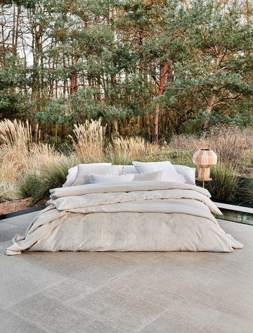 Een bed dat in de buiten ruimte staat. Op het bed ligt een beige dekbedovertrek met een wit patroon en een beige dekbedovertrek met witte strepen. Ook ligt er een beige plaid op het bed