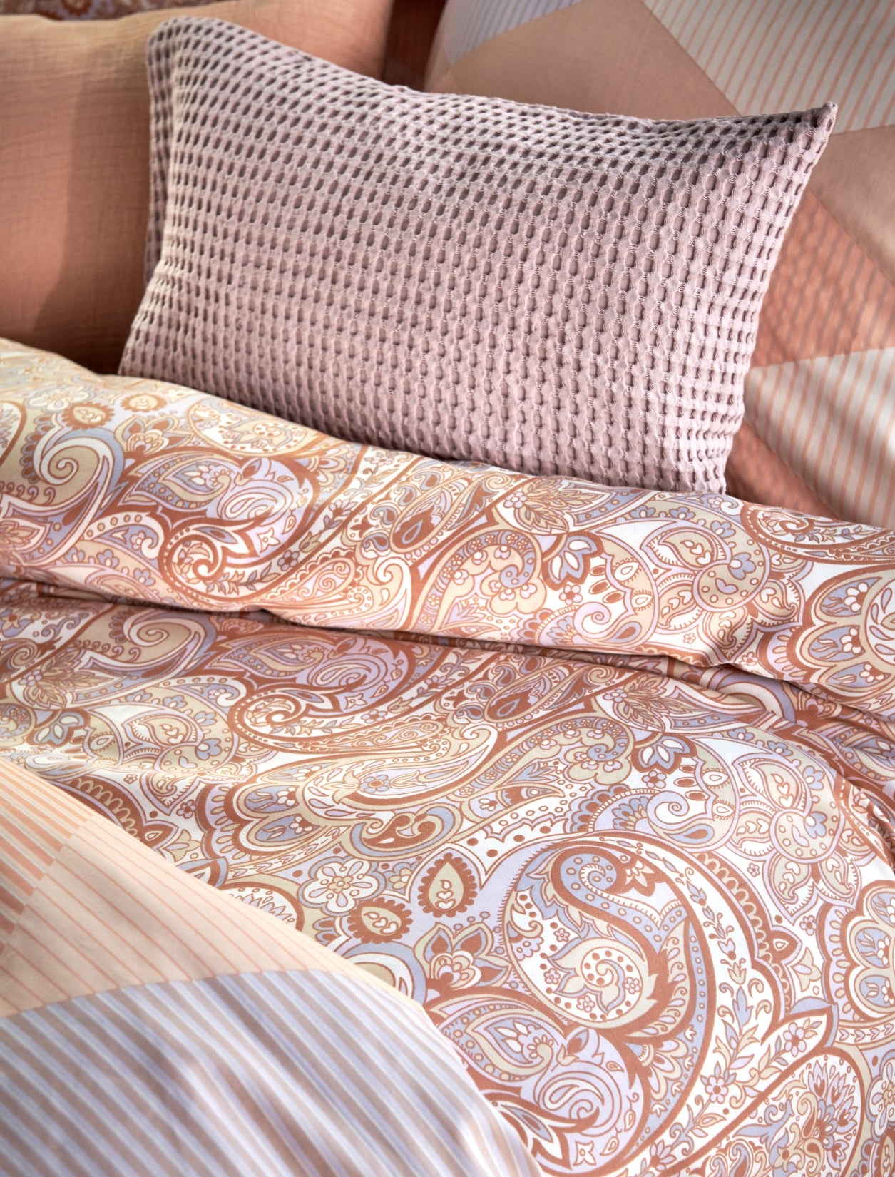detail shot van een dekbedovertrek in een paisley print en een kussen met waffle structuur in lila en pastel oranje kleuren