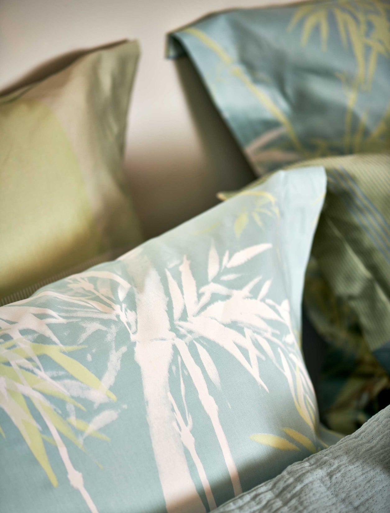 Palm Paradise thyme dekbedovertrek heeft silhouetten van palmbomen in bruin, beige, groen en wit op een lichtblauwe achtergrond. De achterkant van het dekbedovertrek heeft dezelfde print. De kussens hebben aan beide kanten palmbomen.