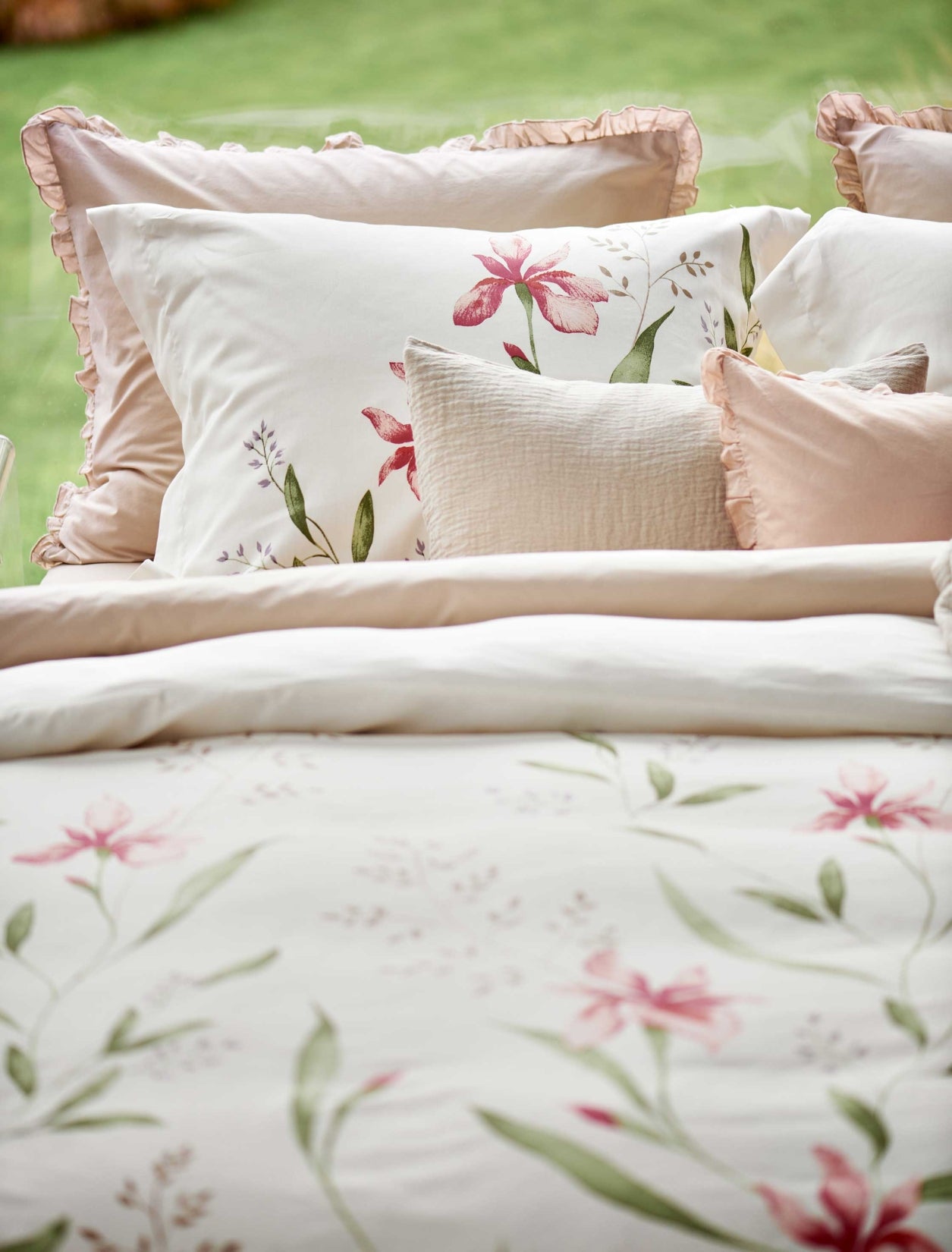 detail shot van een dekbedovertrek met roze bloemen en groene bladeren en een roze ruffle kussens. Op het bed ligt een beige sprei