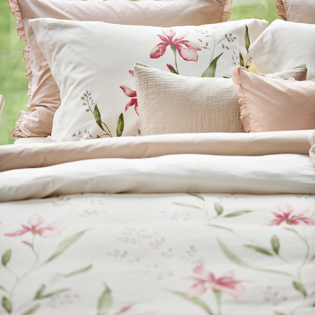 detail shot van een dekbedovertrek met roze bloemen en groene bladeren en een roze ruffle kussens. Op het bed ligt een beige sprei