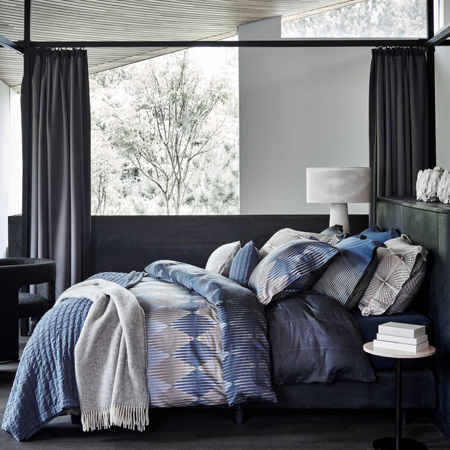 Een bed in een rustige omgeving met een dekbedovertrek met grafische vormen in donkerblauwe kleuren en een grijs plaid