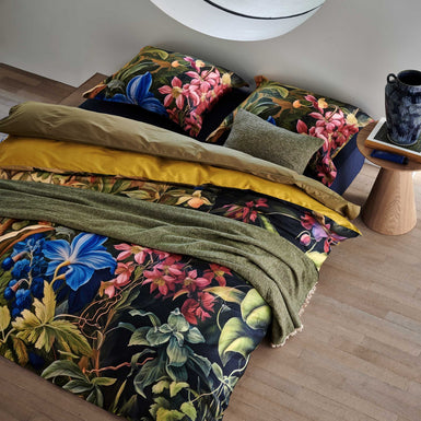 Bed in een rustige omgeving Mystic Garden multi dekbedovertrek heeft imposante bloemen in verschillende tinten roze en blauw, omgeven door grote bladeren en struiken. De achterkant van het dekbedovertrek is effen groen/oker
