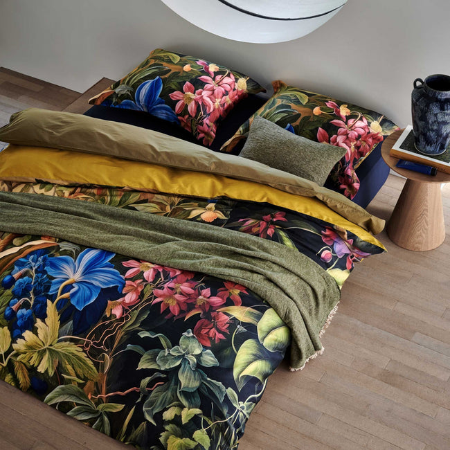 Bed in een rustige omgeving Mystic Garden multi dekbedovertrek heeft imposante bloemen in verschillende tinten roze en blauw, omgeven door grote bladeren en struiken. De achterkant van het dekbedovertrek is effen groen/oker