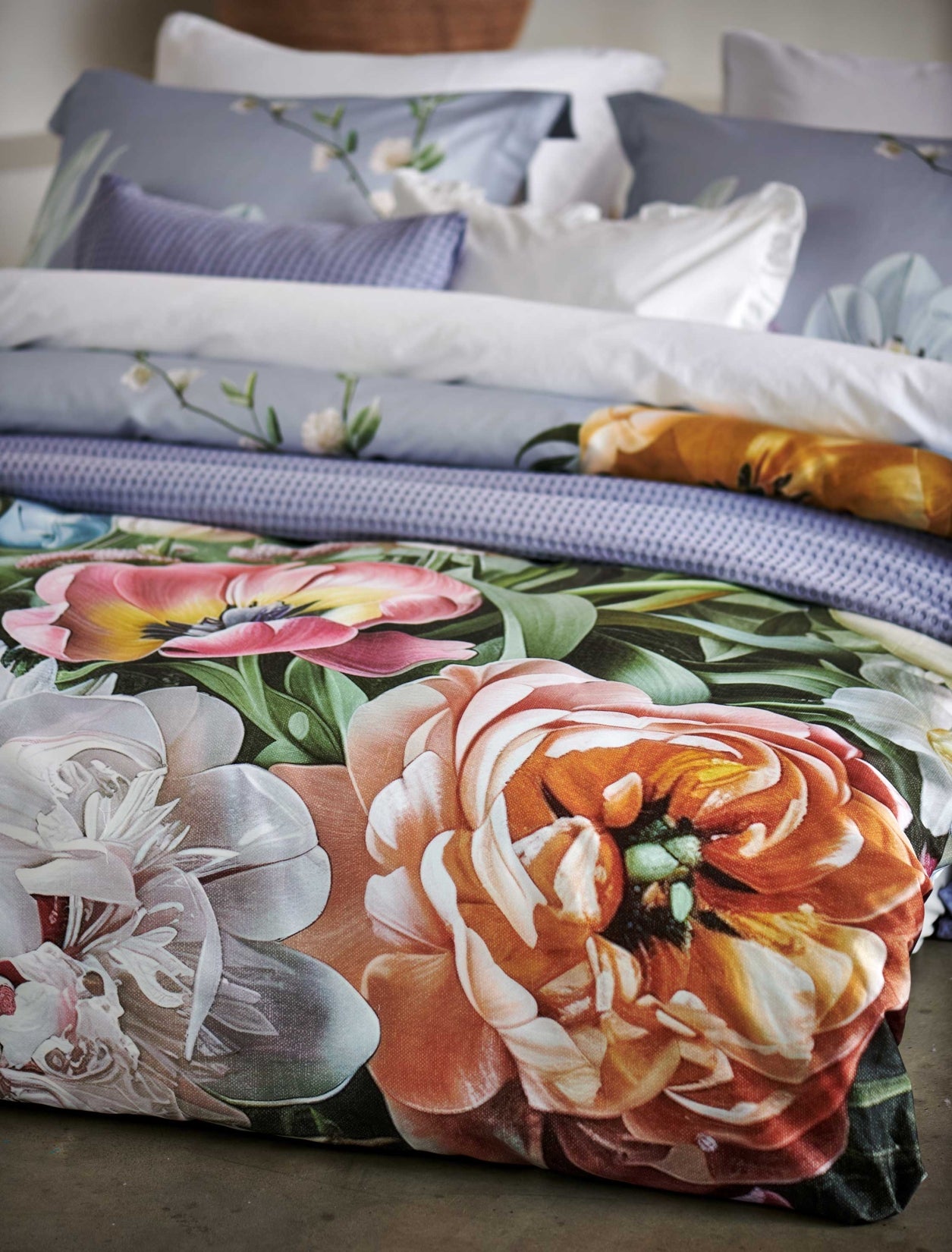 Fleur Festive lavender blue dekbedovertrek heeft hele grote bloemen in lichte pasteltinten in blauw, roze en oranje schakeringen op een blauw / paarse achtergrond. De achterkant van het dekbedovertrek is effen beige. De kussens hebben aan beide kanten een paar bloemen en een bloesemtak.
