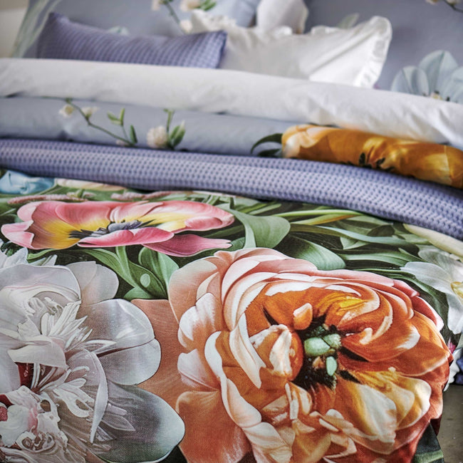 Fleur Festive lavender blue dekbedovertrek heeft hele grote bloemen in lichte pasteltinten in blauw, roze en oranje schakeringen op een blauw / paarse achtergrond. De achterkant van het dekbedovertrek is effen beige. De kussens hebben aan beide kanten een paar bloemen en een bloesemtak.