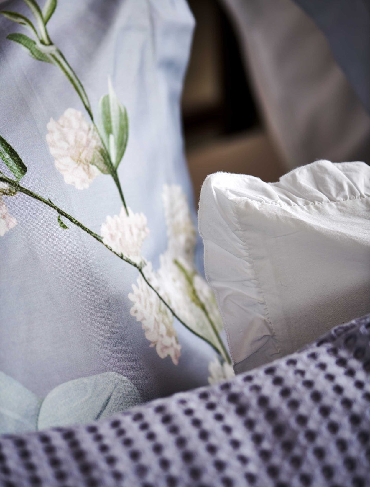 Fleur Festive lavender blue dekbedovertrek heeft hele grote bloemen in lichte pasteltinten in blauw, roze en oranje schakeringen op een blauw / paarse achtergrond. De achterkant van het dekbedovertrek is effen beige. De kussens hebben aan beide kanten een paar bloemen en een bloesemtak.